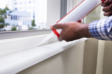 A person uses a caulking gun to apply sealant to a white window frame.