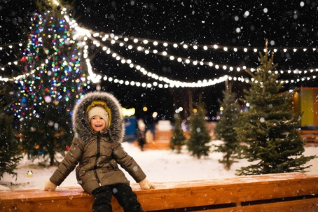 A smiling child in a warm winter coat sits outside on a snowy night